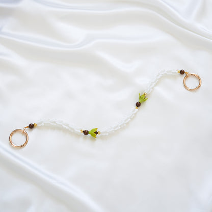 Delicate bag lei featuring white crown flower and two green pakalana flower beads and dark circle beads intertwined with gold rings on each end on a white fabric background