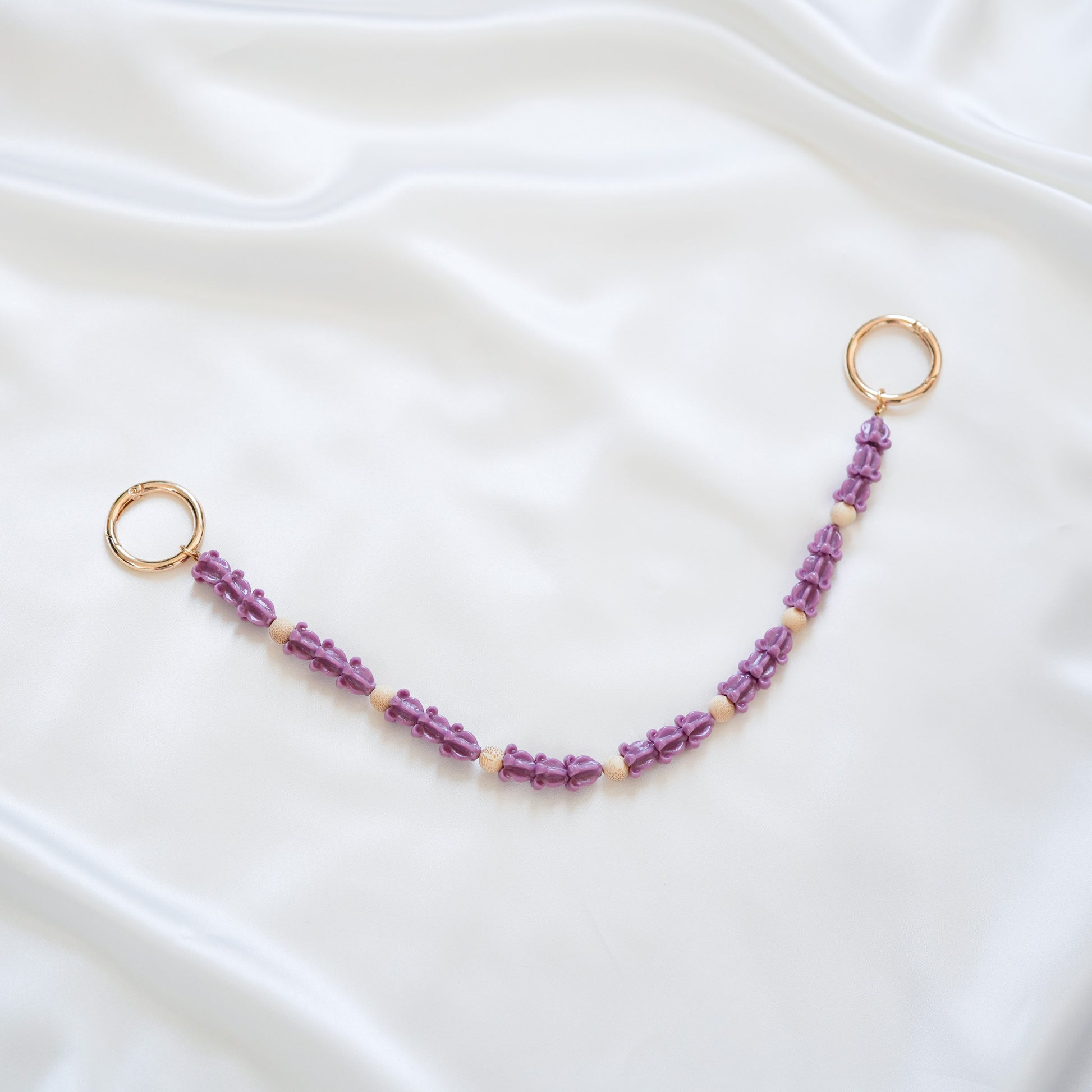 Delicate bag lei featuring dark purple crown flower in sets of three separated by light tan beads with gold rings on each end on a white fabric background