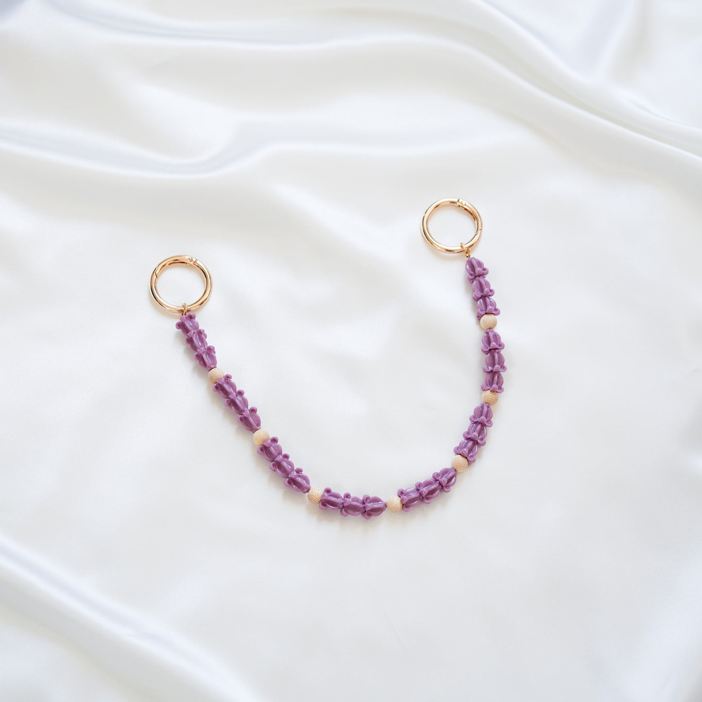 Delicate bag lei featuring dark purple crown flower in sets of three separated by light tan beads with gold rings on each end on a white fabric background