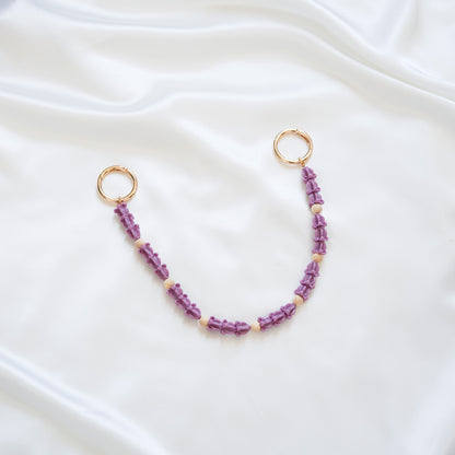 Delicate bag lei featuring dark purple crown flower in sets of three separated by light tan beads with gold rings on each end on a white fabric background