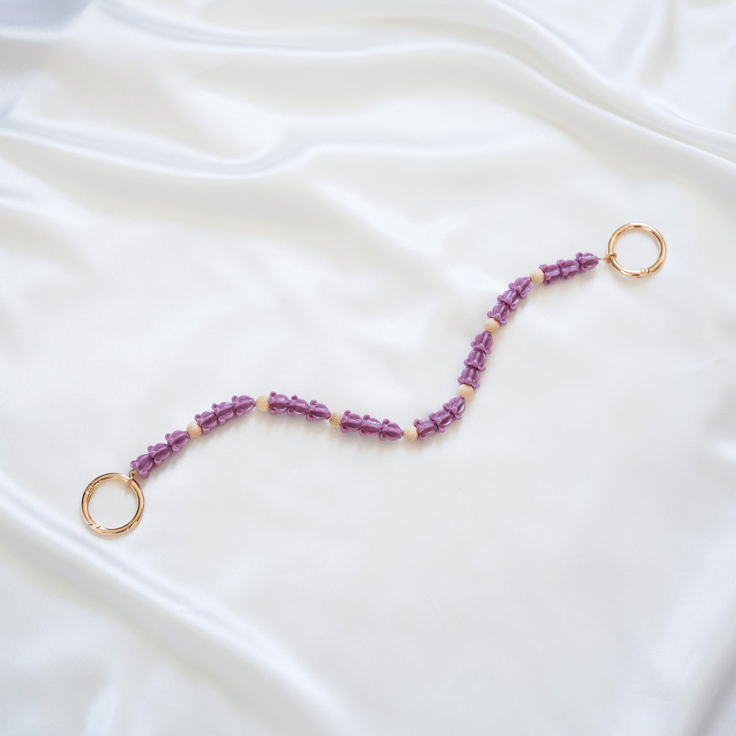 Delicate bag lei featuring dark purple crown flower in sets of three separated by light tan beads with gold rings on each end on a white fabric background