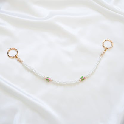 Delicate bag lei featuring white crown flower and two pink roses with gold rings on each end on a white fabric background