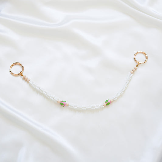 Delicate bag lei featuring white crown flower and two pink roses with gold rings on each end on a white fabric background