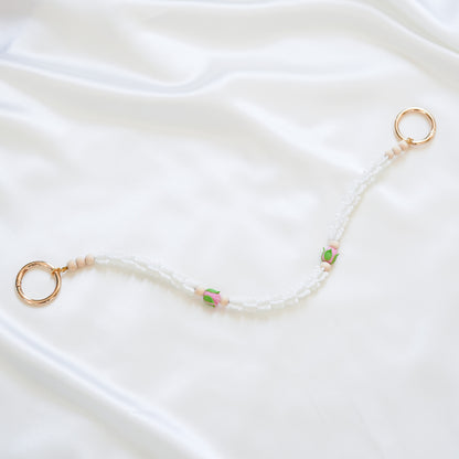 Delicate bag lei featuring white crown flower and two pink rose beads with gold rings on each end on a white fabric background