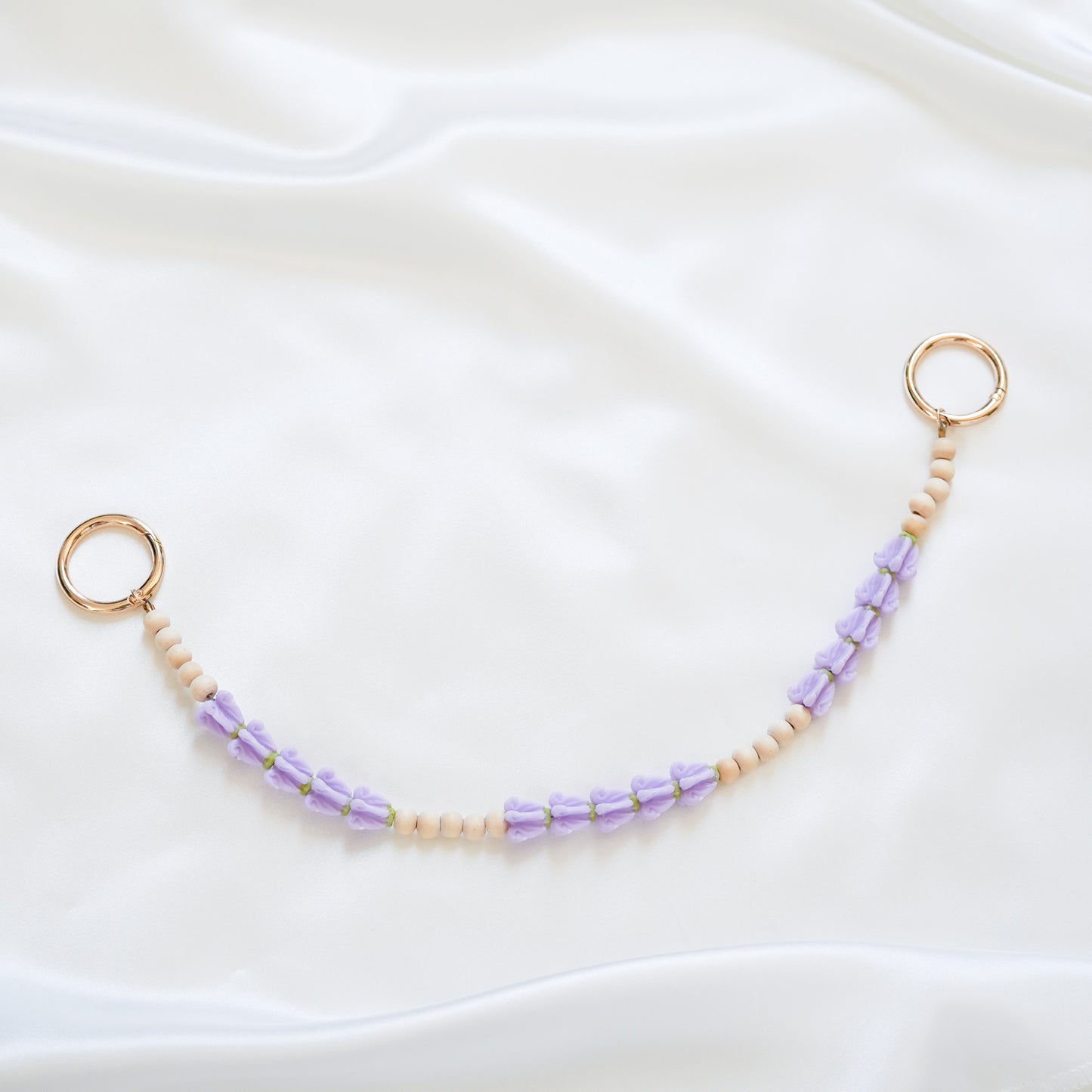 Delicate bag lei featuring purple crown flower and tan beads alternating in sets of five with gold rings on each end on a white fabric background