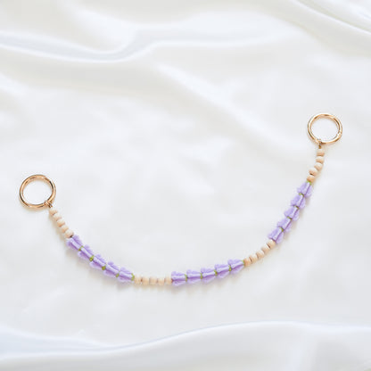 Delicate bag lei featuring purple crown flower and tan beads alternating in sets of five with gold rings on each end on a white fabric background