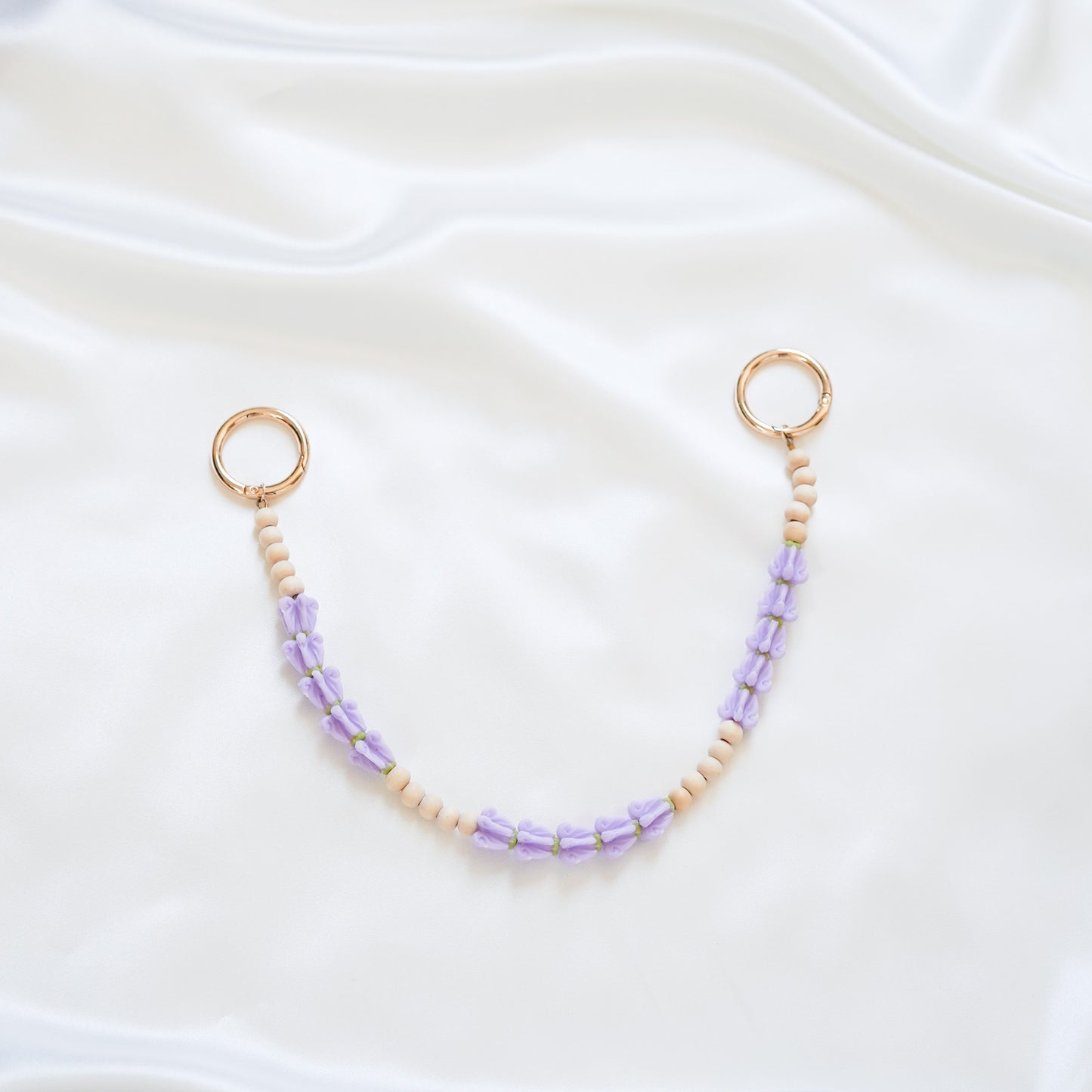 Delicate bag lei featuring purple crown flower and tan beads alternating in sets of five with gold rings on each end on a white fabric background