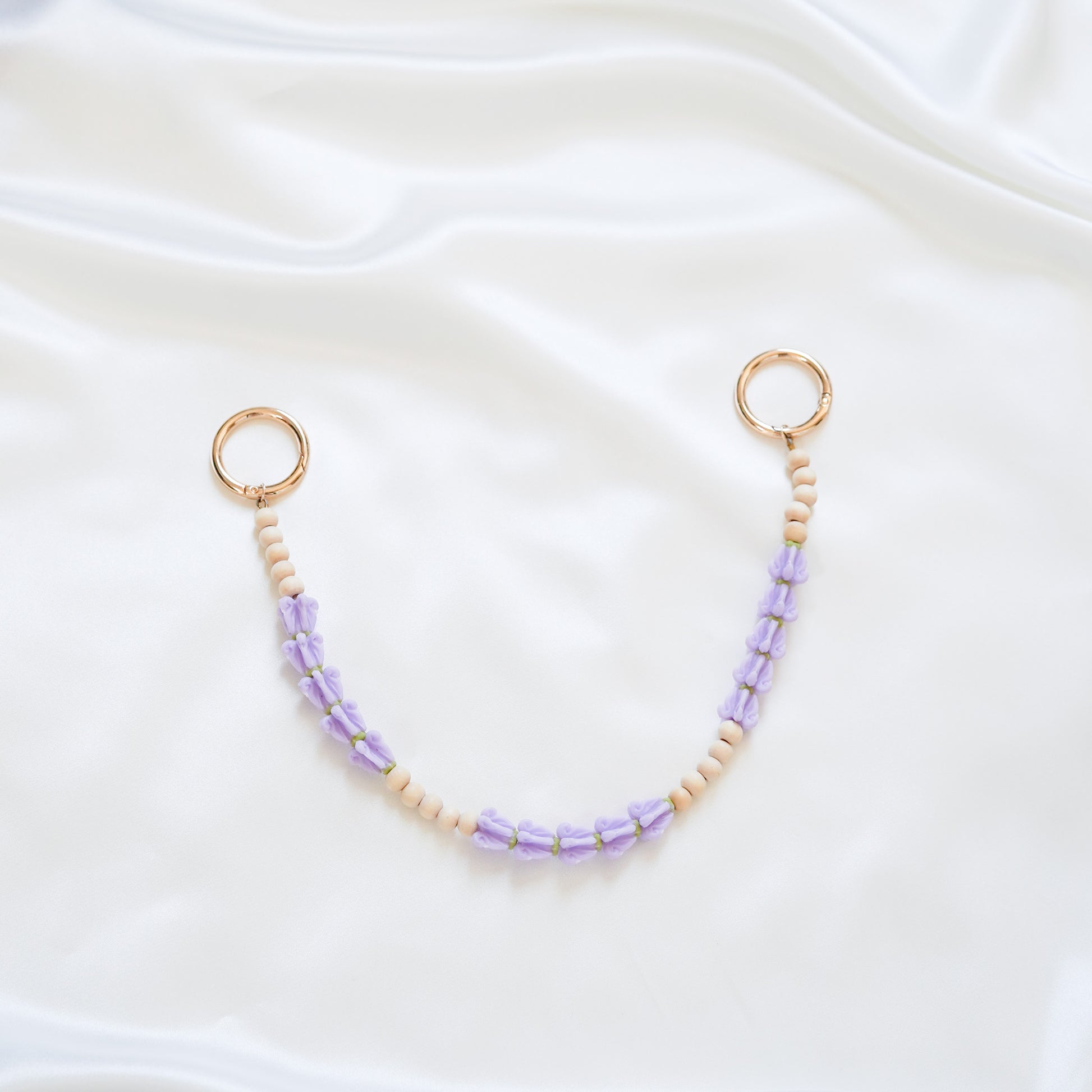 Delicate bag lei featuring purple crown flower and tan beads alternating in sets of five with gold rings on each end on a white fabric background