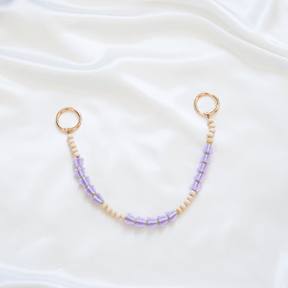 Delicate bag lei featuring purple crown flower and tan beads alternating in sets of five with gold rings on each end on a white fabric background