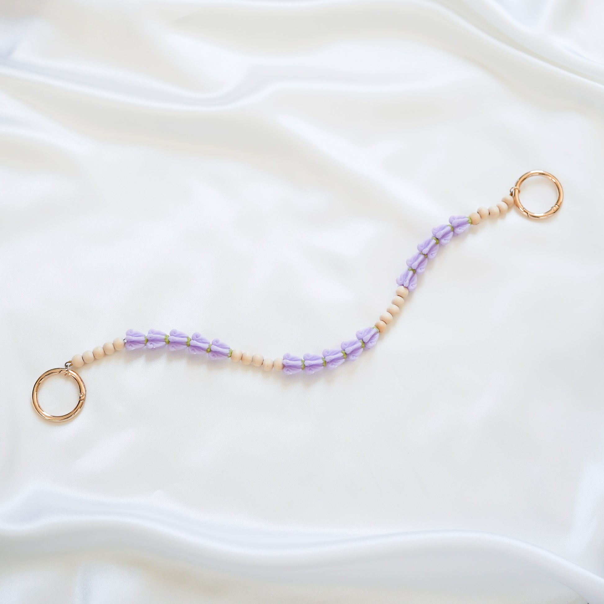 Delicate bag lei featuring purple crown flower and tan beads alternating in sets of five with gold rings on each end on a white fabric background