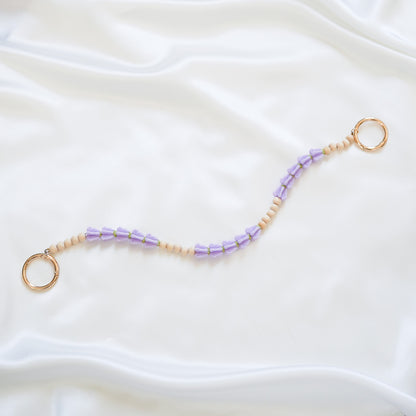 Delicate bag lei featuring purple crown flower and tan beads alternating in sets of five with gold rings on each end on a white fabric background