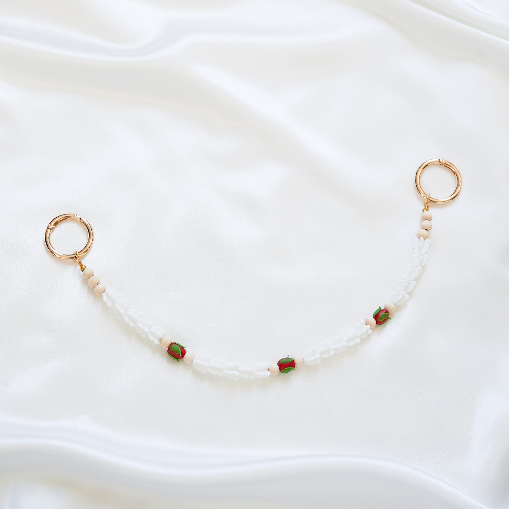 Delicate bag lei featuring white crown flower and three red rose beads with gold rings on each end on a white fabric background