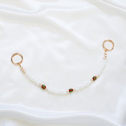 Delicate bag lei featuring white crown flower and three red rose beads with gold rings on each end on a white fabric background