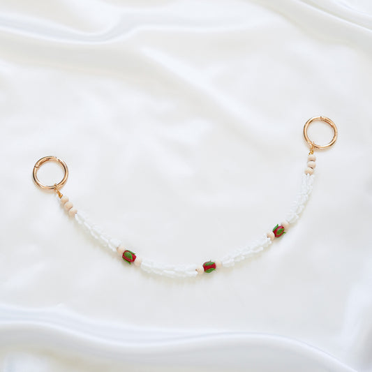 Delicate bag lei featuring white crown flower and three red rose beads with gold rings on each end on a white fabric background