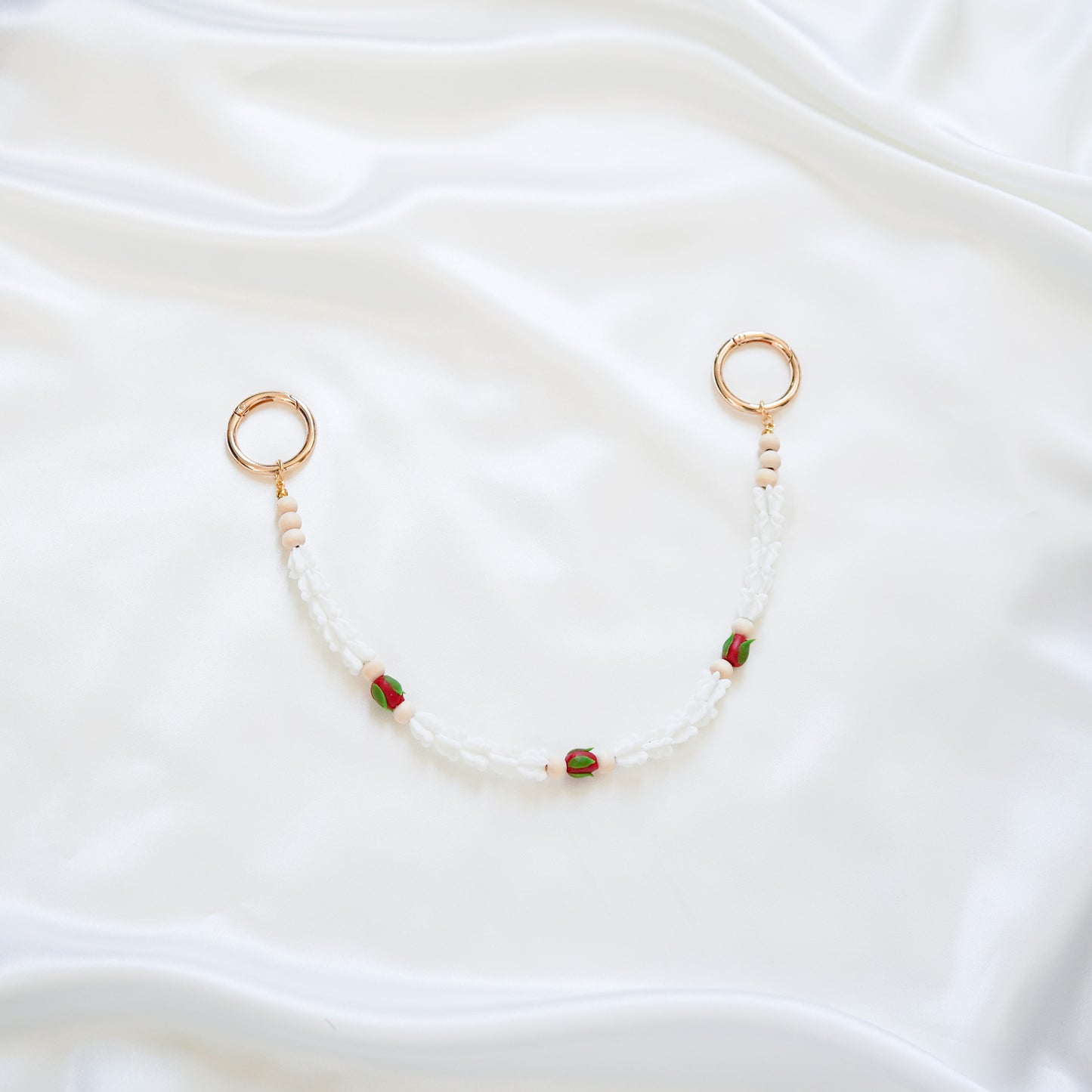 Delicate bag lei featuring white crown flower and three red rose beads with gold rings on each end on a white fabric background