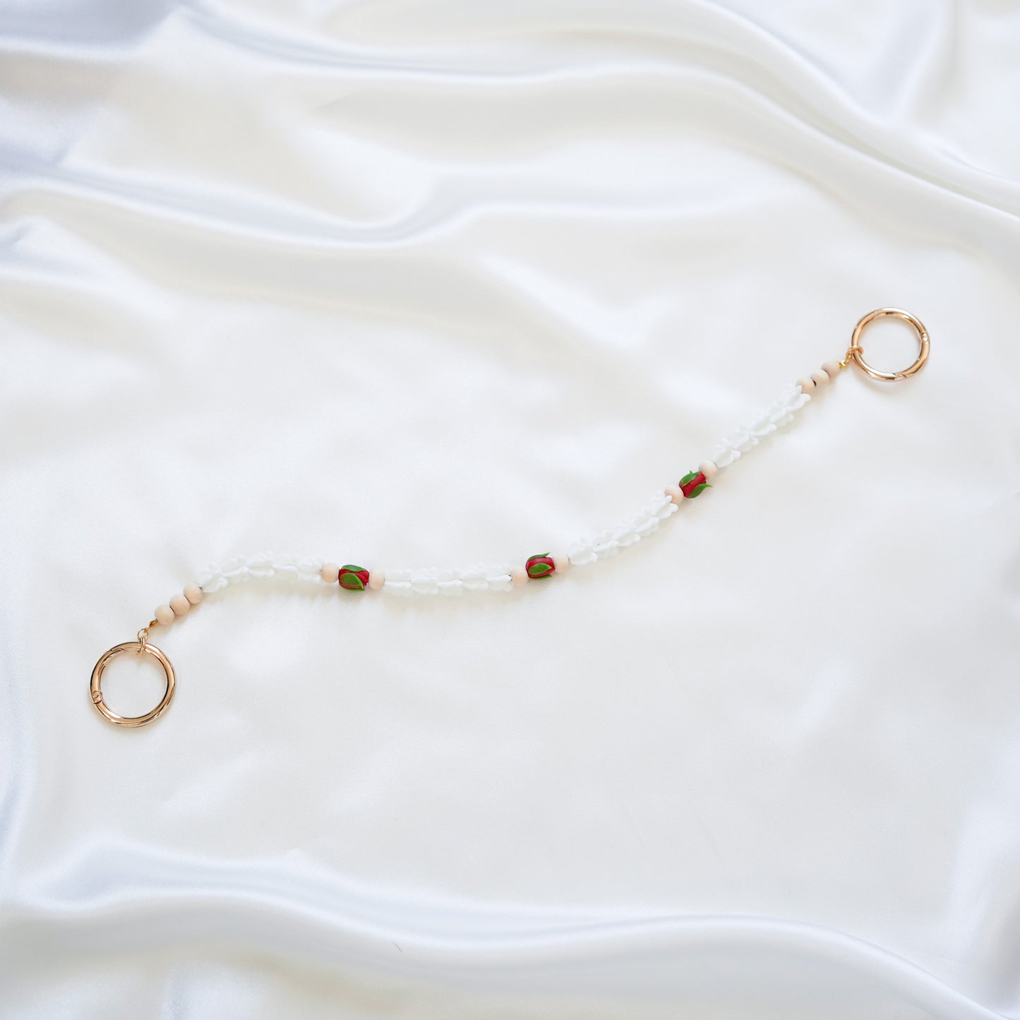 Delicate bag lei featuring white crown flower and three red rose beads with gold rings on each end on a white fabric background