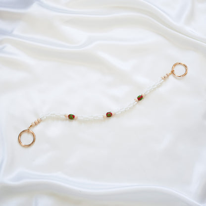 Delicate bag lei featuring white crown flower and three red rose beads with gold rings on each end on a white fabric background