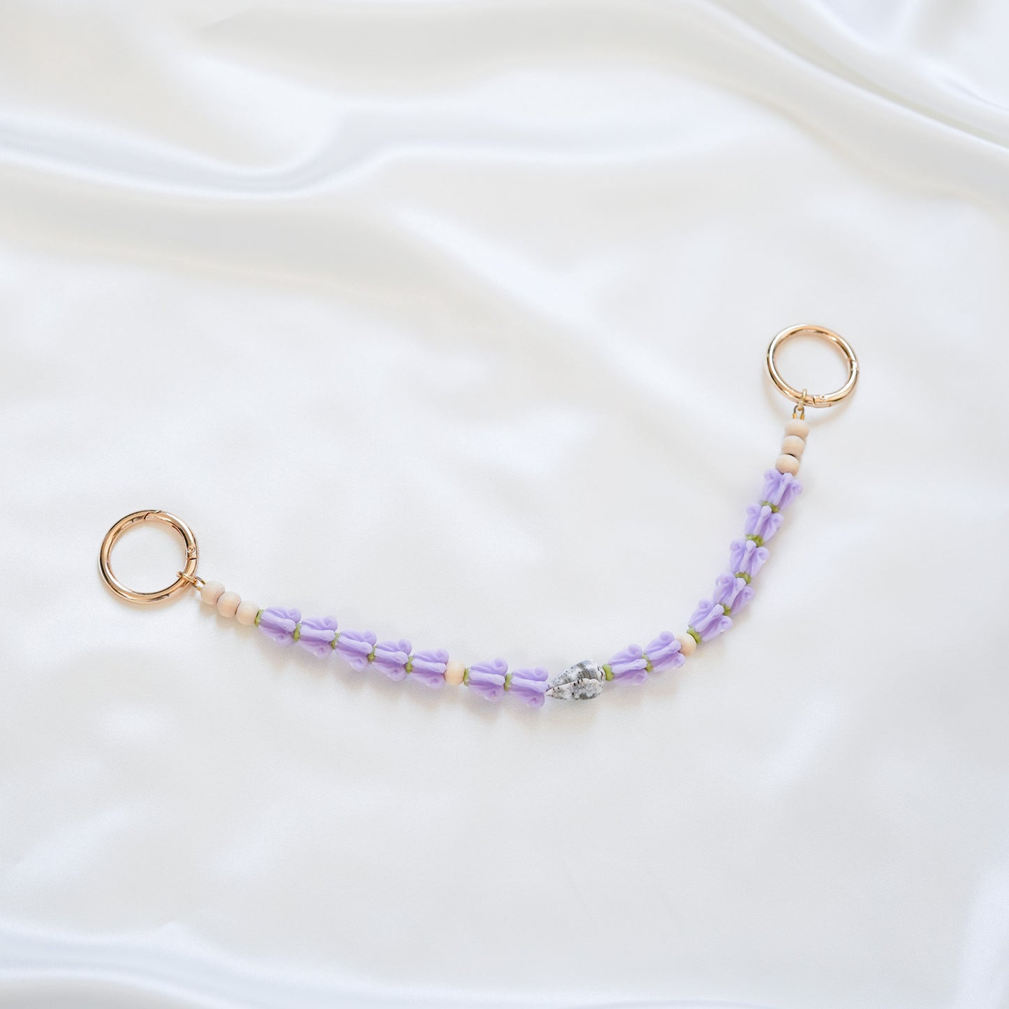 Delicate bag lei featuring purple crown flower and a patterned shell in the middle with gold rings on each end on a white fabric background