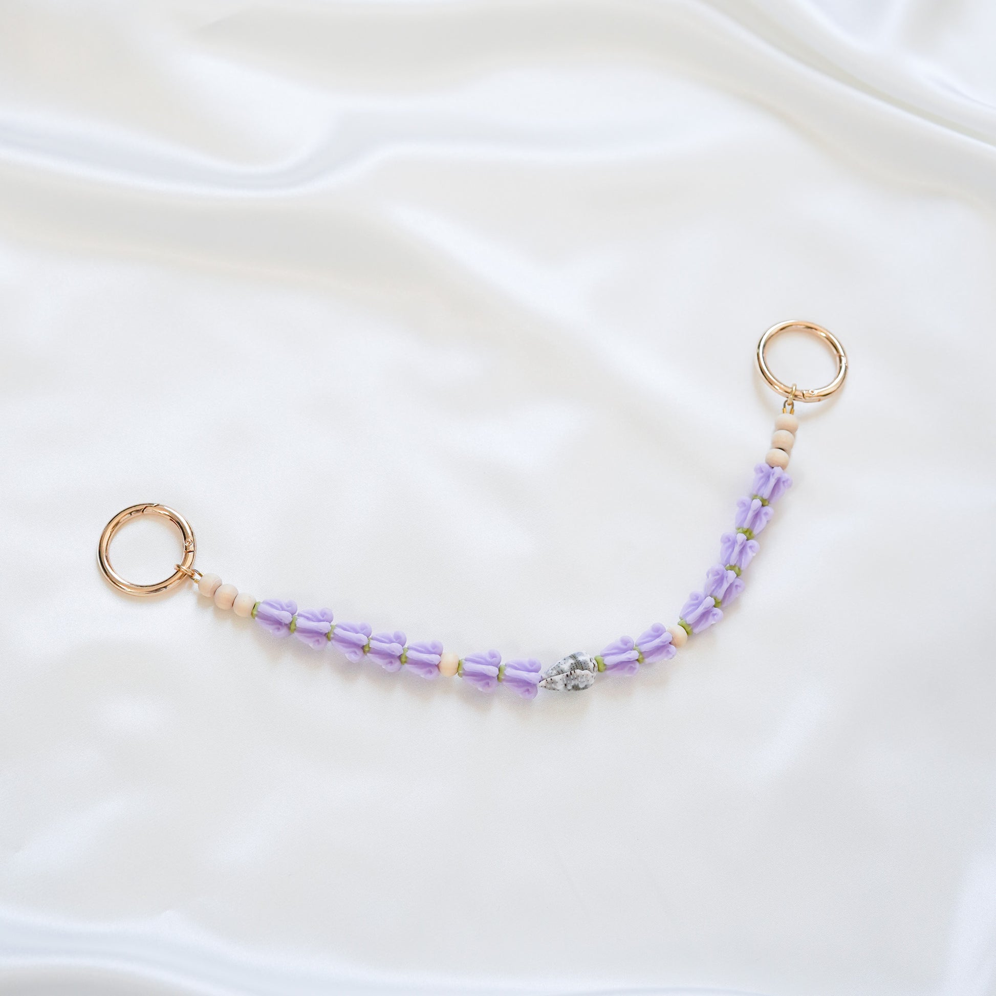 Delicate bag lei featuring purple crown flower and a patterned shell in the middle with gold rings on each end on a white fabric background