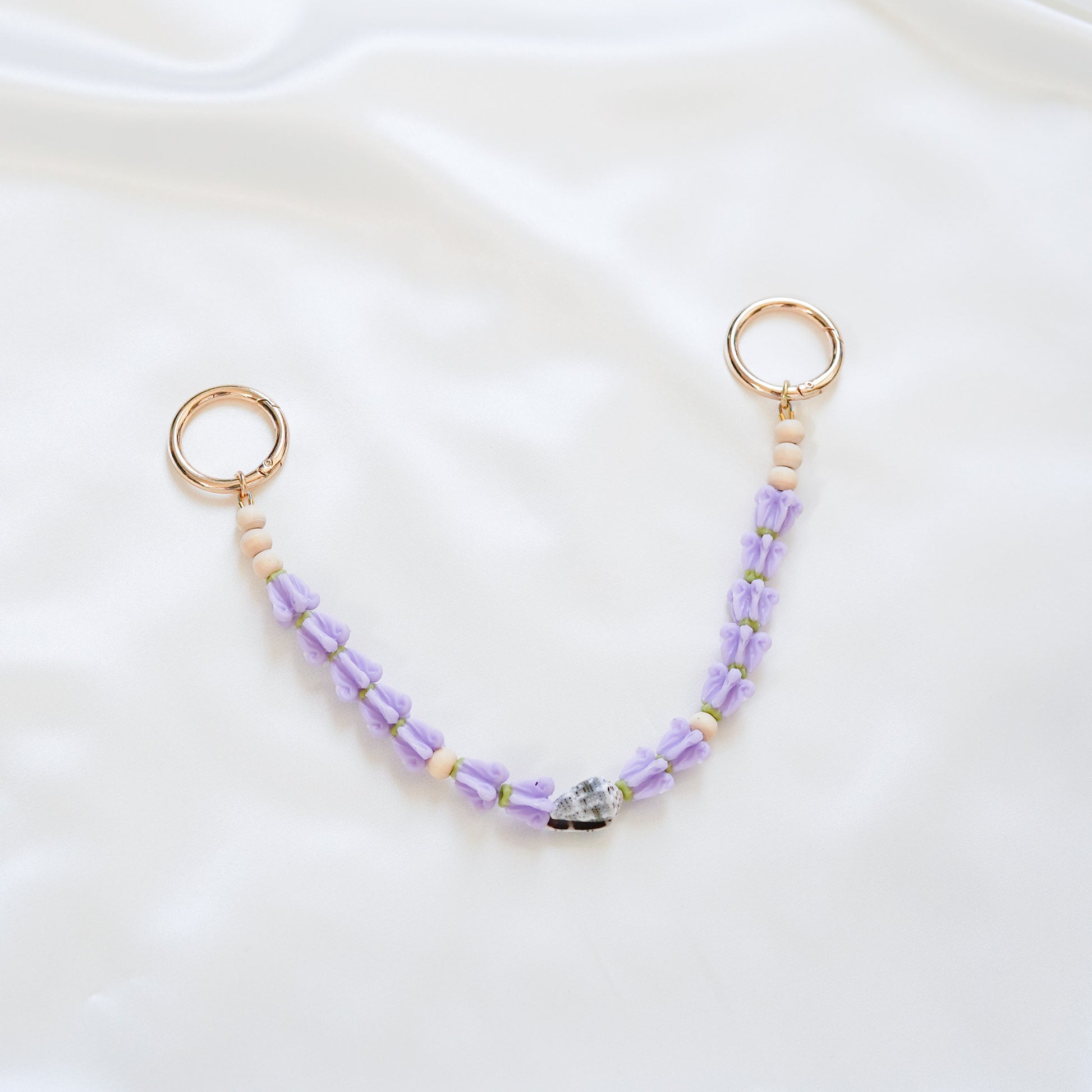 Delicate bag lei featuring purple crown flower and a patterned shell in the middle with gold rings on each end on a white fabric background