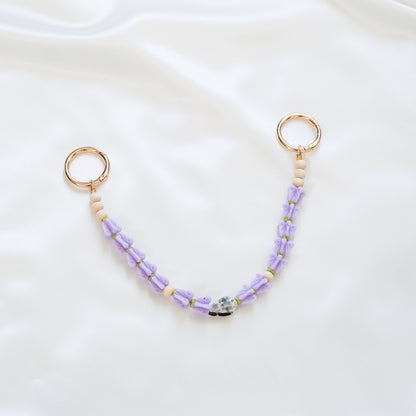 Delicate bag lei featuring purple crown flower and a patterned shell in the middle with gold rings on each end on a white fabric background