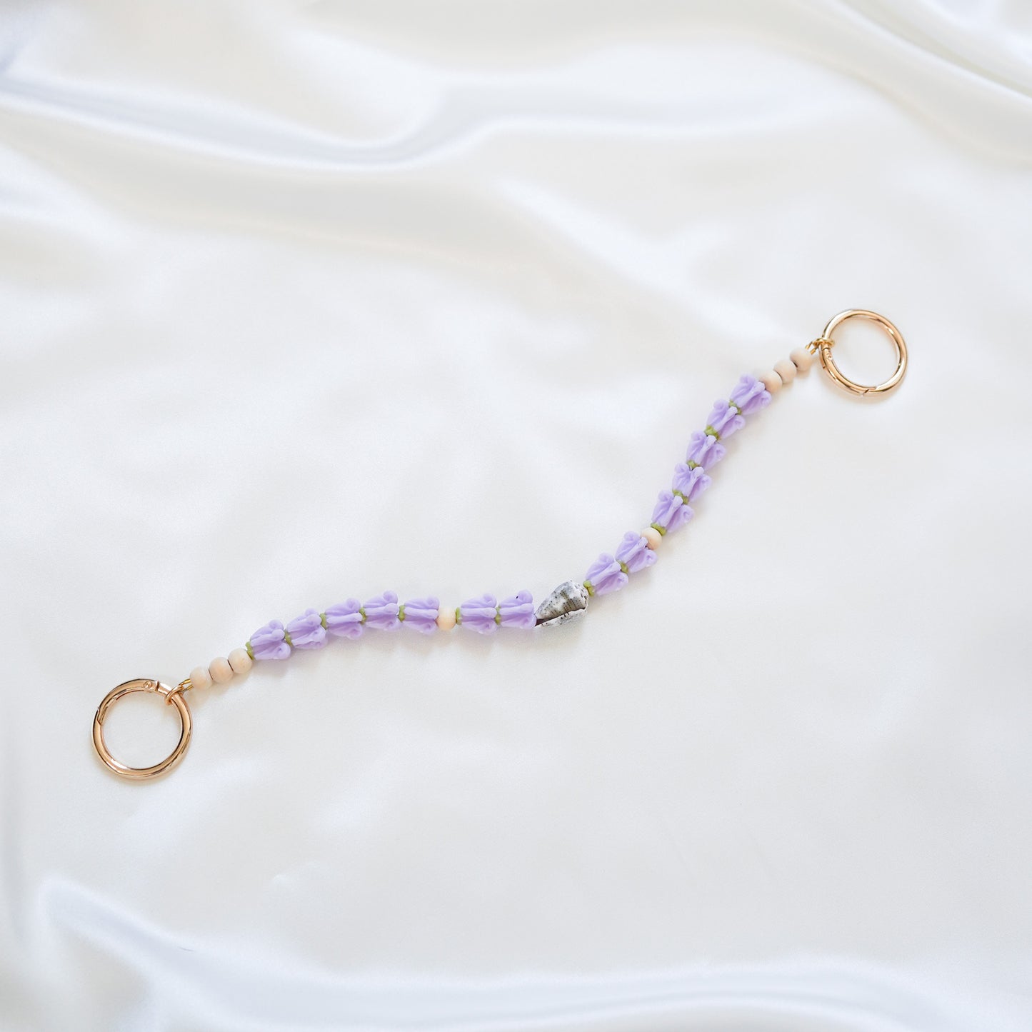 Delicate bag lei featuring purple crown flower and a patterned shell in the middle with gold rings on each end on a white fabric background
