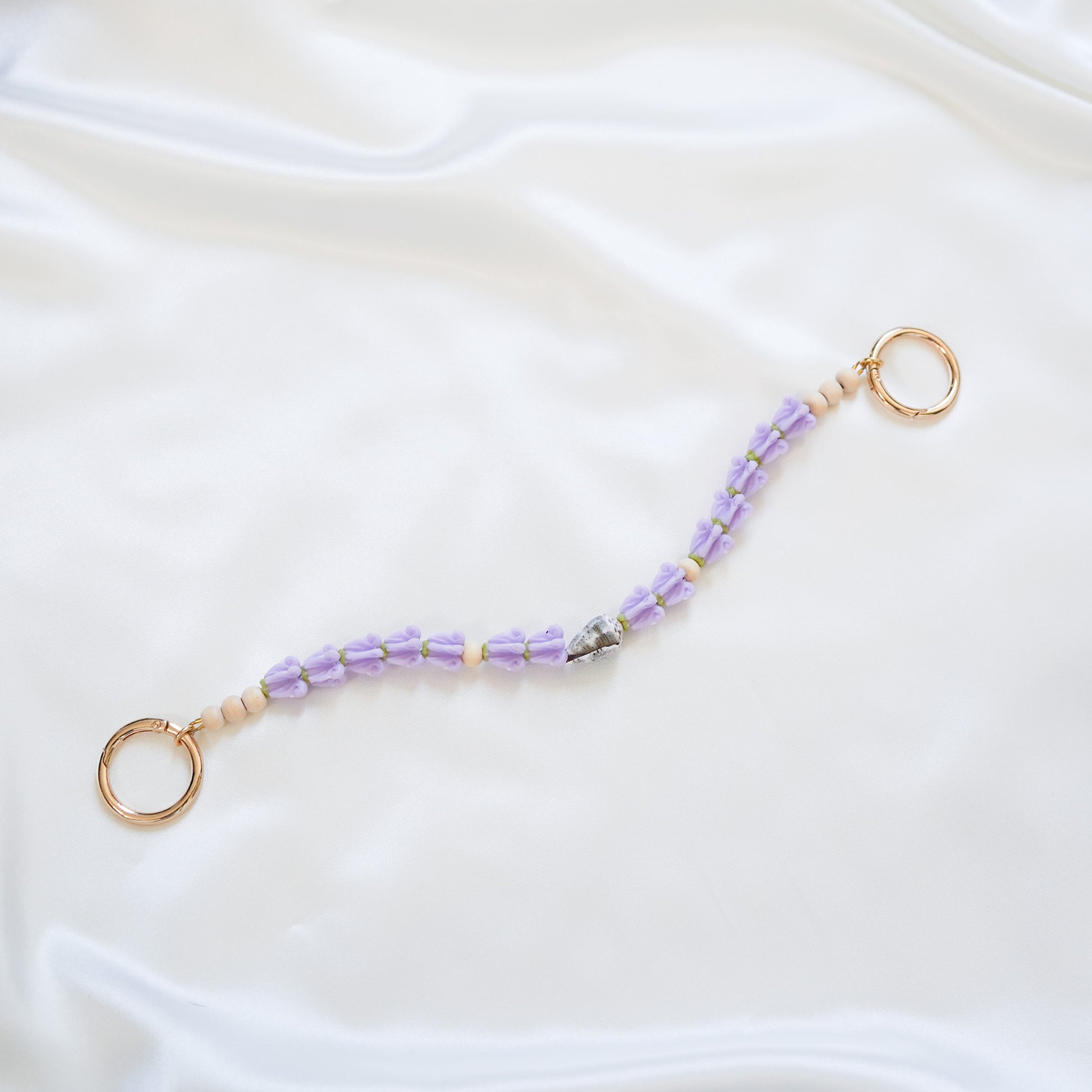 Delicate bag lei featuring purple crown flower and a patterned shell in the middle with gold rings on each end on a white fabric background