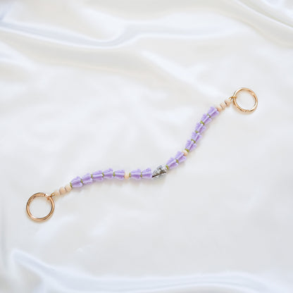 Delicate bag lei featuring purple crown flower and a patterned shell in the middle with gold rings on each end on a white fabric background
