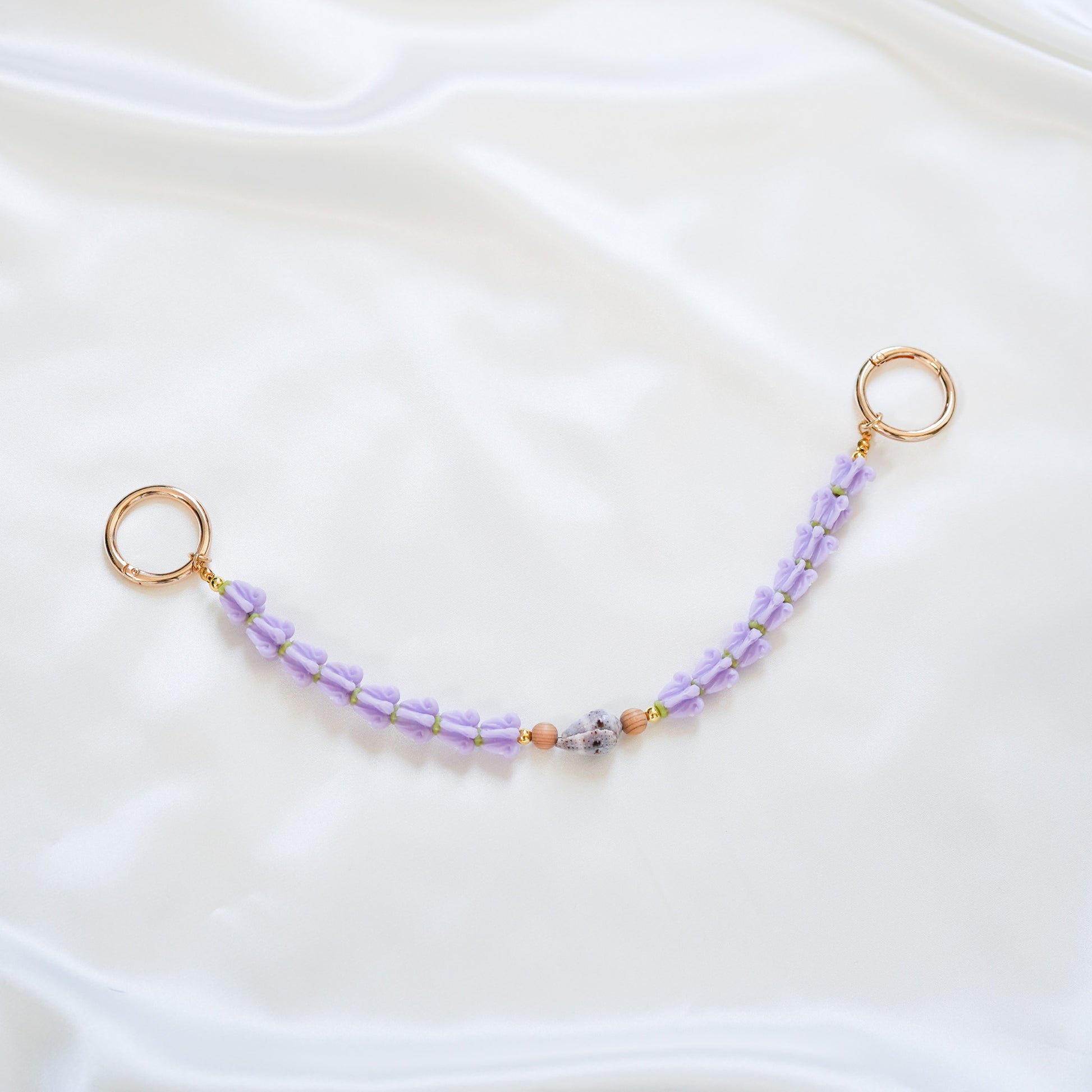 Delicate bag lei featuring purple crown flower and a patterned shell in the middle with tan beads around shell with gold rings on each end on a white fabric background