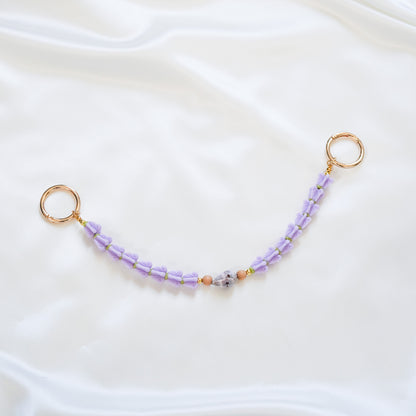 Delicate bag lei featuring purple crown flower and a patterned shell in the middle with tan beads around shell with gold rings on each end on a white fabric background