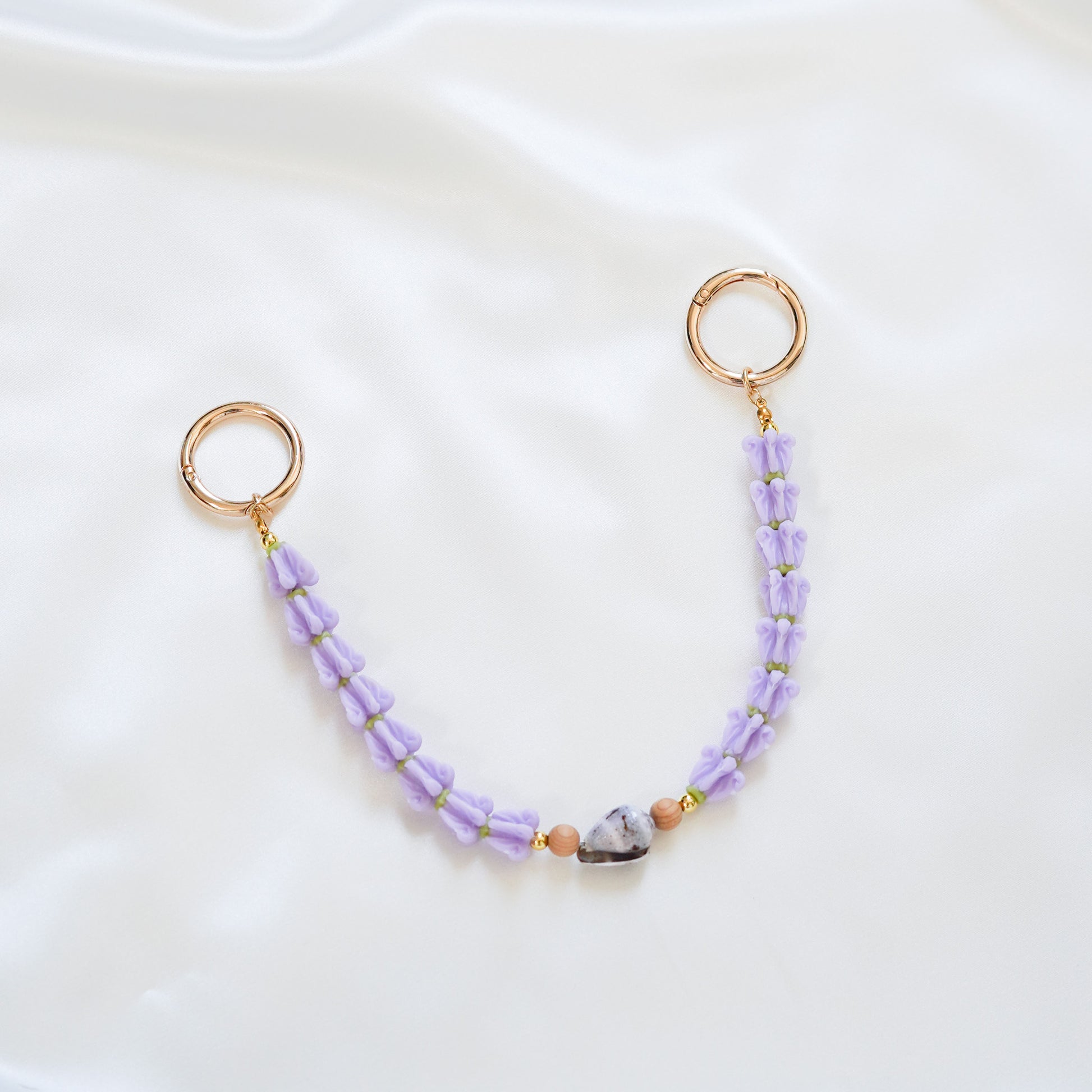 Delicate bag lei featuring purple crown flower and a patterned shell in the middle with tan beads around shell with gold rings on each end on a white fabric background