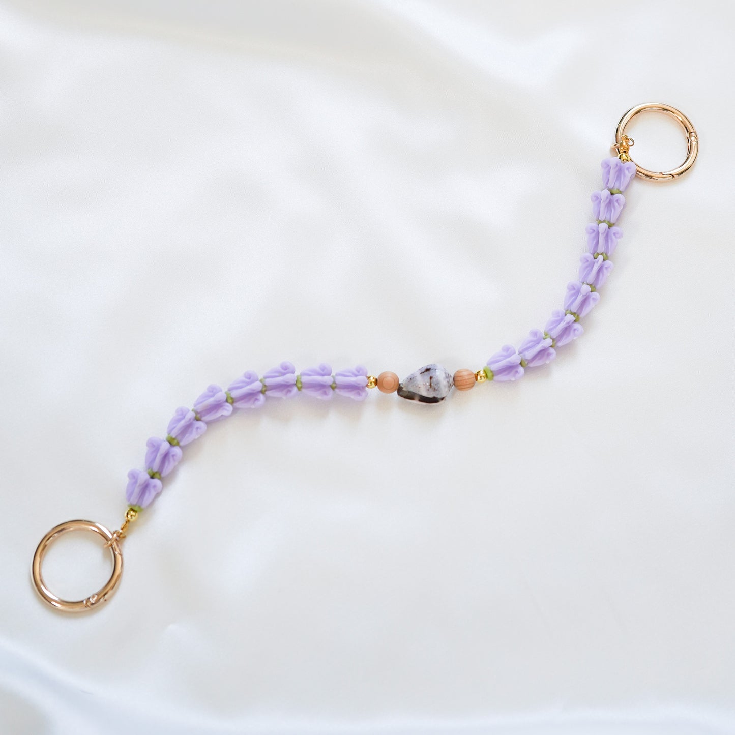 Delicate bag lei featuring purple crown flower and a patterned shell in the middle with tan beads around shell with gold rings on each end on a white fabric background
