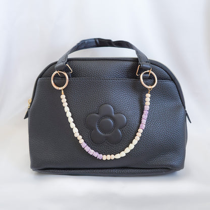 Black handbag with a floral design and beaded bag lei on the handle on a white background