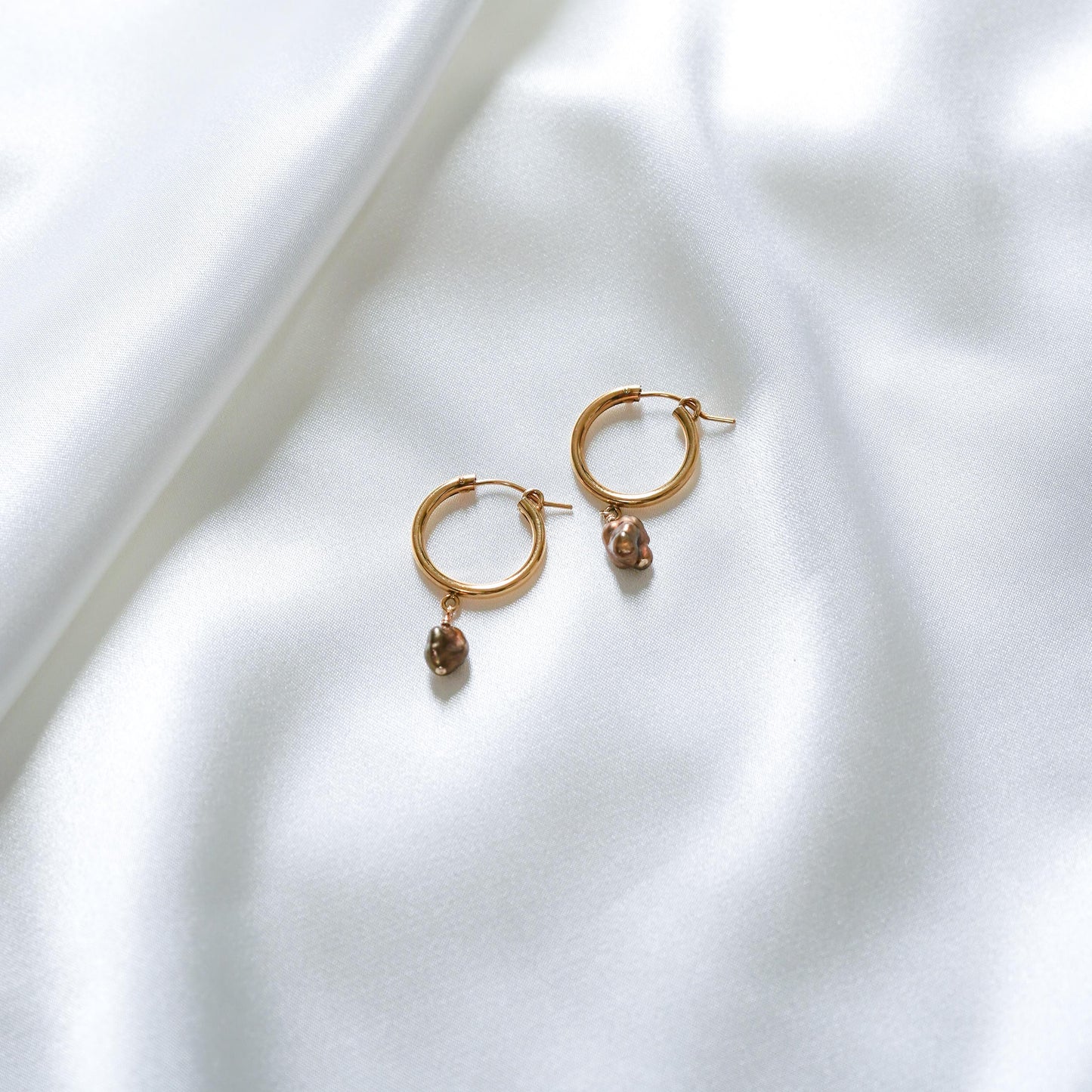Gold hoop earrings with small chocolate keshi charms on a white fabric background