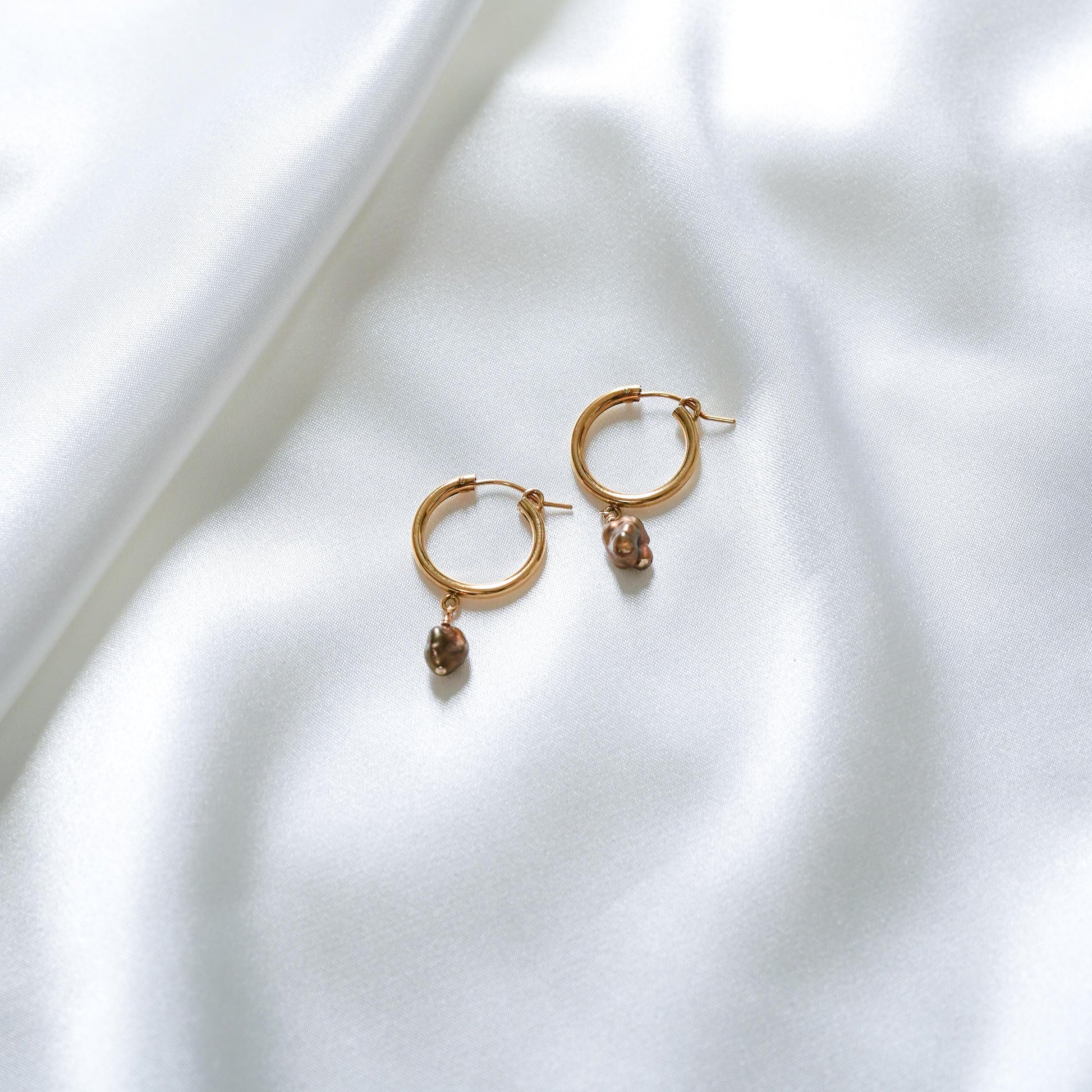 Gold hoop earrings with small chocolate keshi charms on a white fabric background