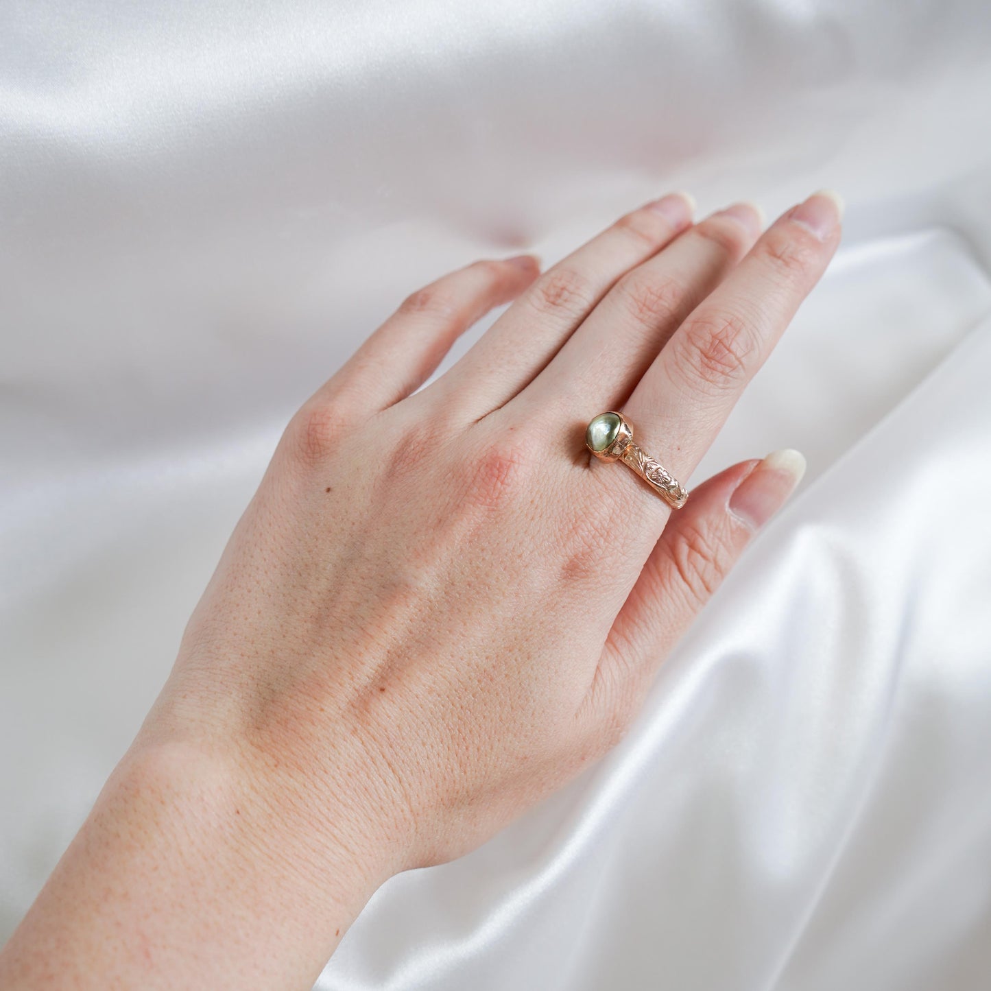 Hand wearing a ring with a green keshi pearl on a white satin background