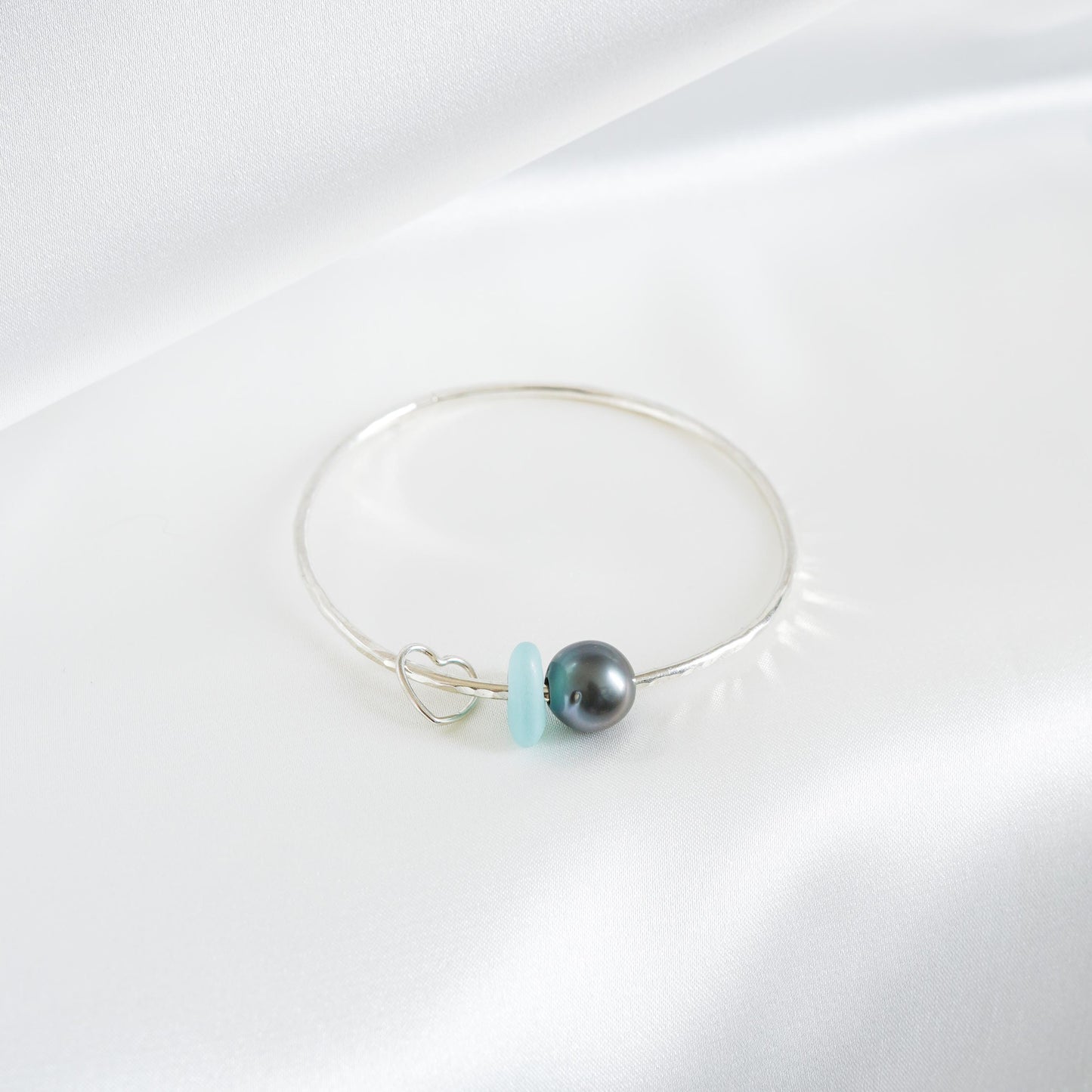 Silver bracelet with a tahitian pearl, light blue sea glass and heart charm on a white background