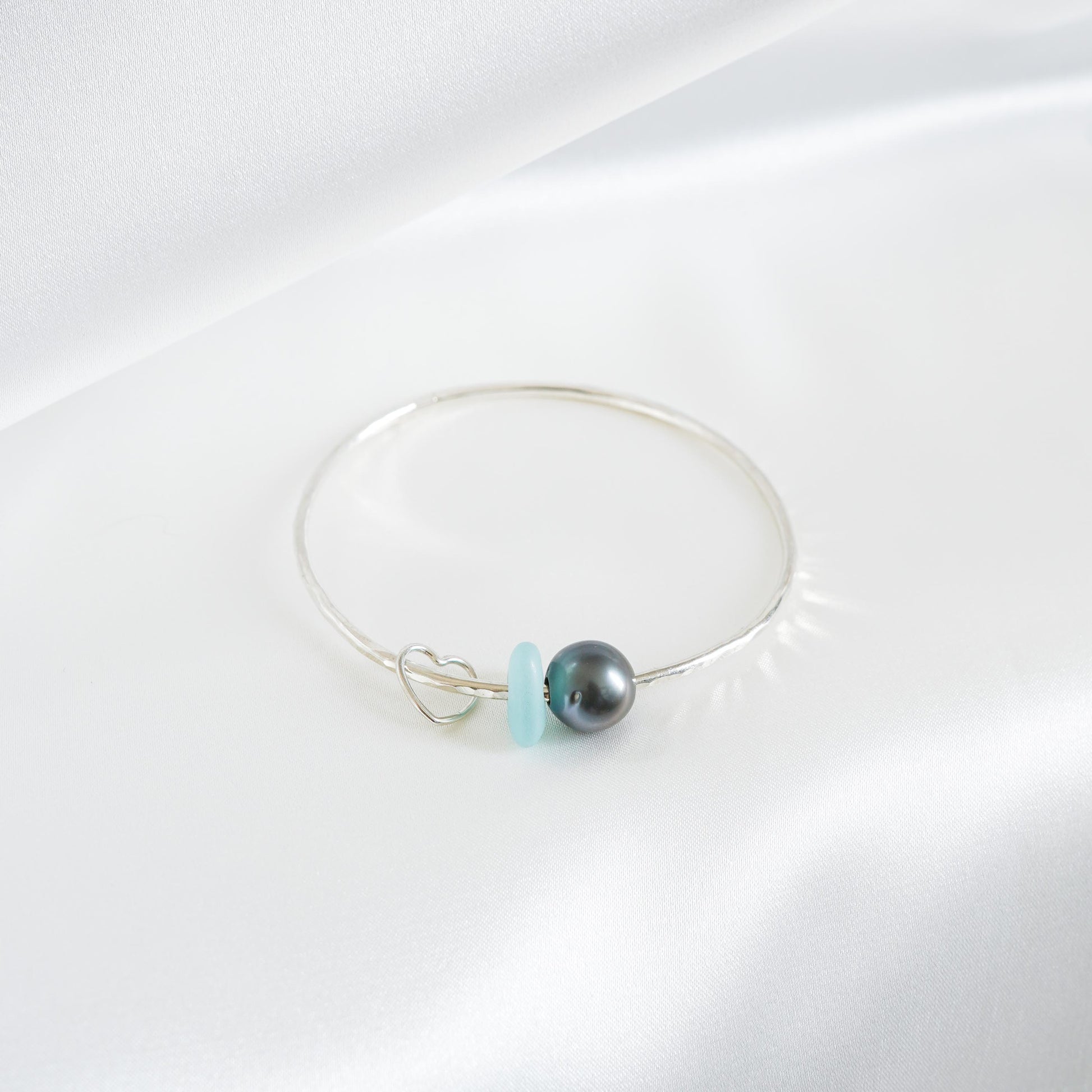 Silver bracelet with a tahitian pearl, light blue sea glass and heart charm on a white background