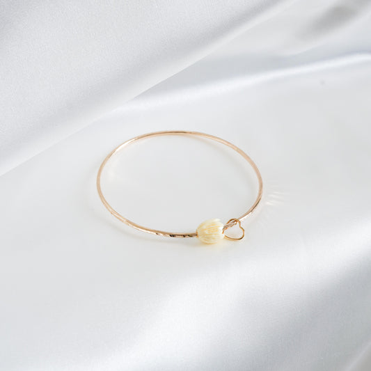 Gold bracelet with a tan pikake flower bead with heart charm on a white fabric background