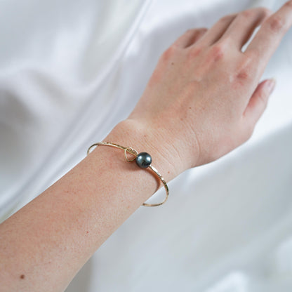 Hand wearing a gold bangle with tahitian pearl and gold heart charm