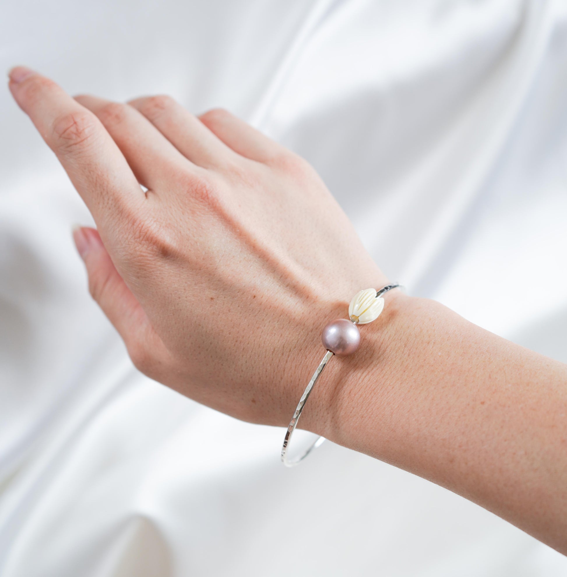 Silver bangle with pink pearl and tan pikake bead on a wrist against white satin background