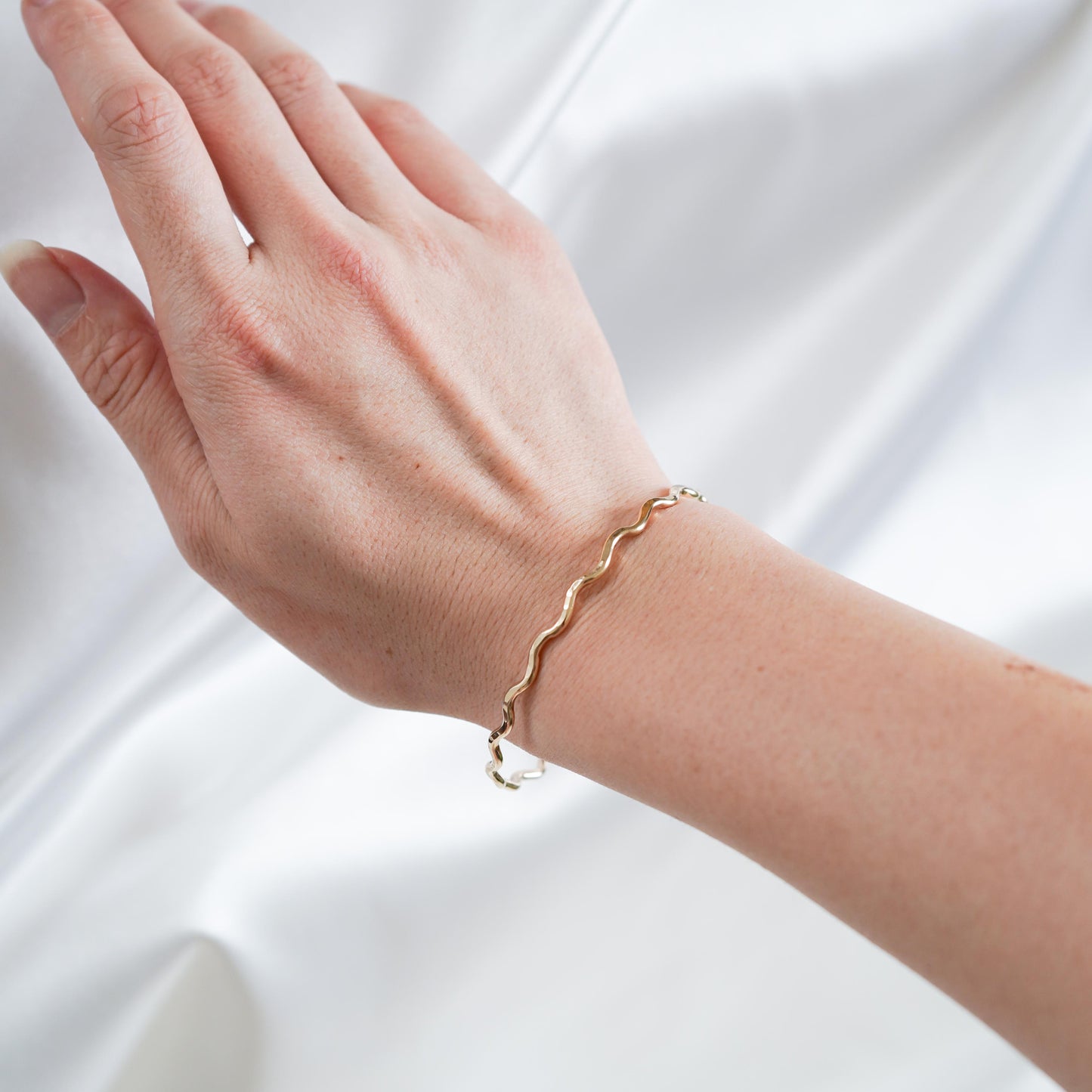 gold wavy bangle on wrist against white satin fabric
