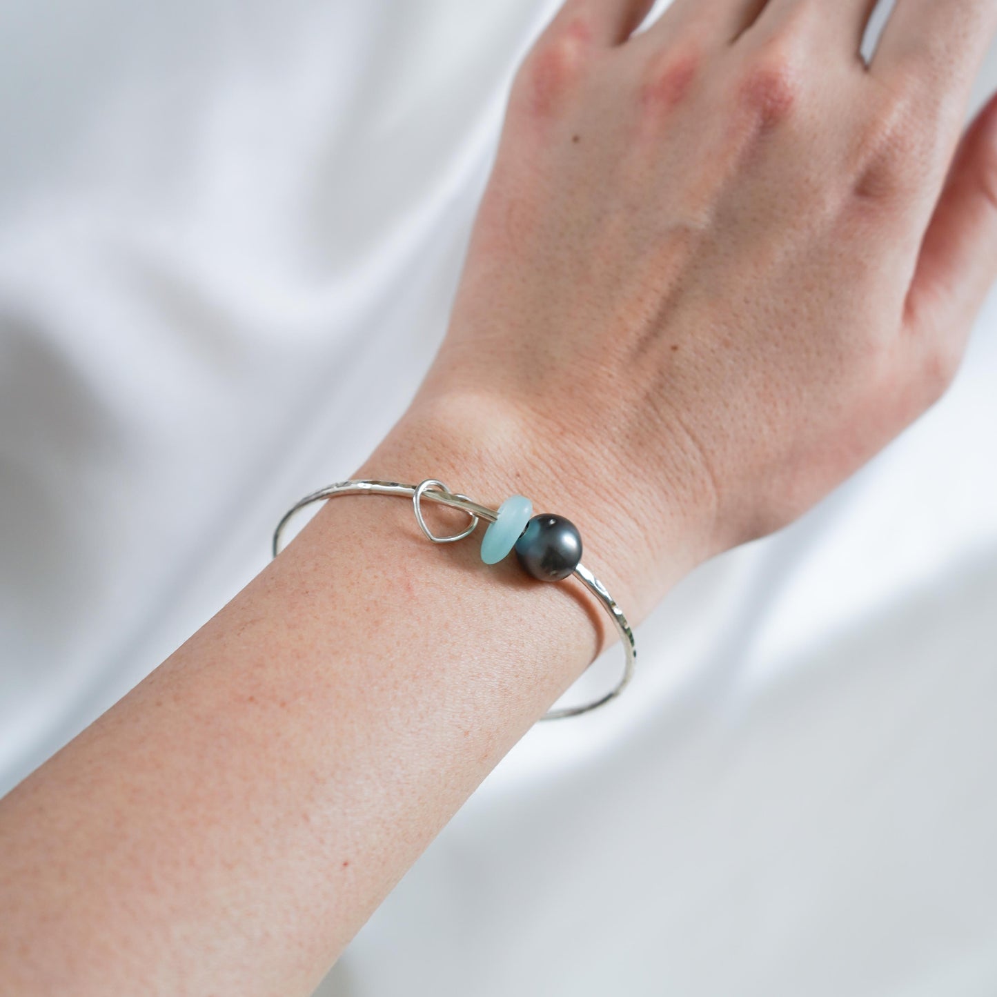 Wrist wearing a silver bangle with a tahitian pearl, blue sea glass and heart charm