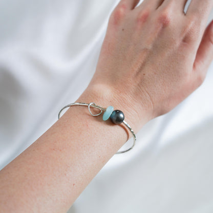 Wrist wearing a silver bangle with a tahitian pearl, blue sea glass and heart charm
