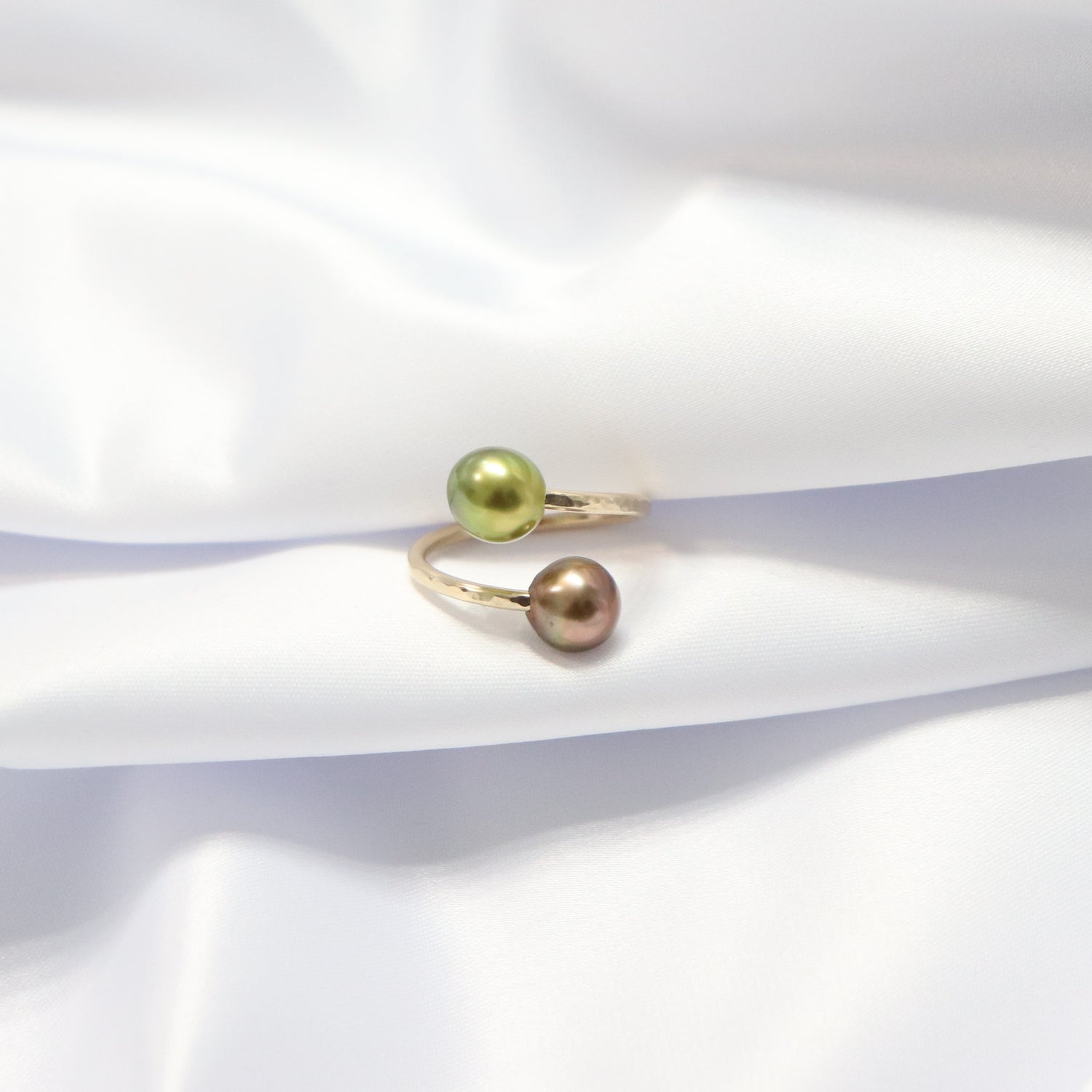 Gold ring with one green and one brown pearl on a white fabric background