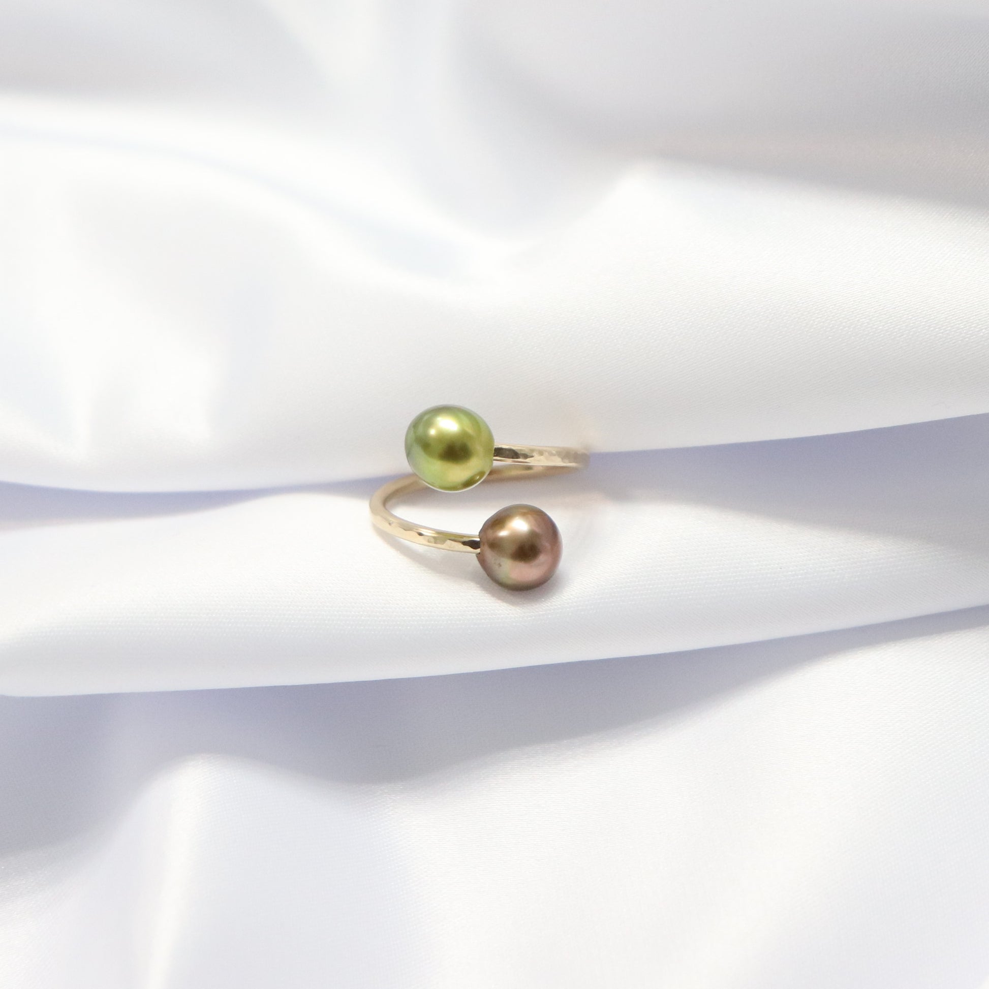 Gold ring with one green and one brown pearl on a white fabric background